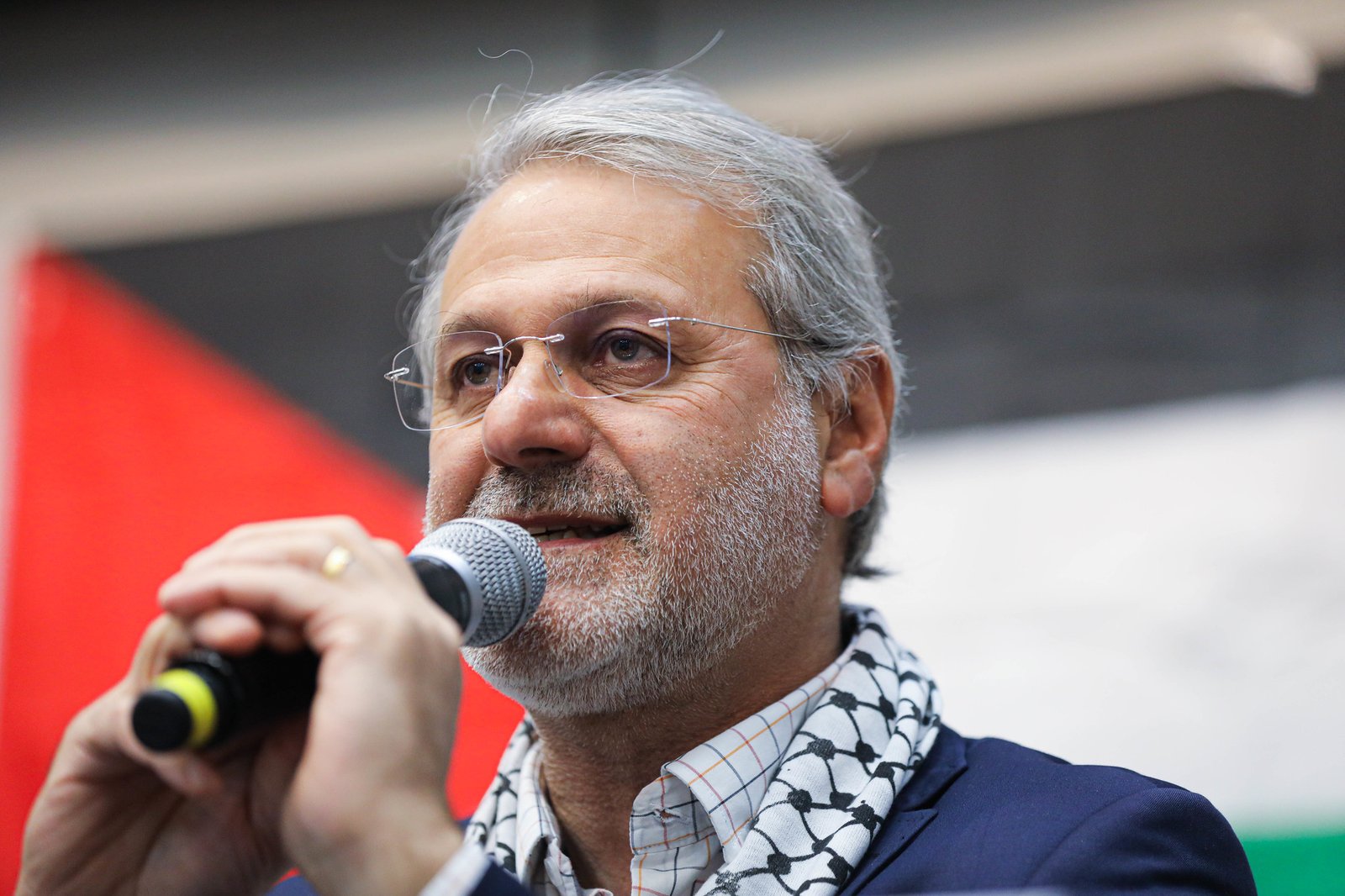 Ualid Hussein Ali Mohd Rabah discursando ao microfone, usando kufiya, com a bandeira da Palestina ao fundo